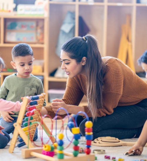 Learning Through Play A female Kindergarten teacher of Middle Eastern decent, sits on the floor with students as they play with various toys and engage in different activities. They are each dressed casually as they learn through their play.